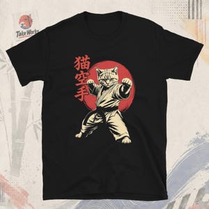 Könnte beinhalten: Ein schwarzes Kurzarm-T-Shirt mit einer Grafik eines Katzenmotivs in einem Kampfkunst-Anzug mit schwarzem Gürtel. Die Katze befindet sich in einer Kampfhaltung vor einem großen roten Kreis. Rote japanische Schriftzeichen sind links neben der Katze.
