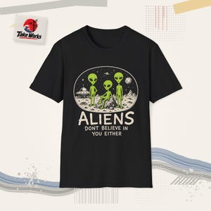 May include: A black short-sleeved t-shirt featuring a graphic of three green aliens on a moon surface with a flying saucer. Below the illustration, the text reads "ALIENS DONT BELIEVE IN YOU EITHER" in a bold, off-white, sans-serif font.