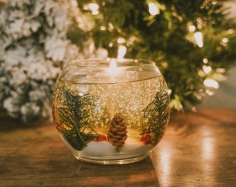 May include: A clear glass jar candle with a pine cone, greenery, and red berries inside. The candle is lit and the wax is melted, creating a warm glow.