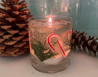 May include: A clear glass jar candle with a red and white candy cane, green pine sprigs, and white glitter inside. The candle is lit and the flame is visible.
