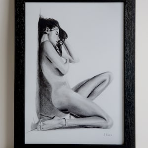 Può includere: Un disegno a carboncino in bianco e nero di una donna nuda seduta con la testa appoggiata sul braccio. Il disegno è incorniciato in una cornice nera.