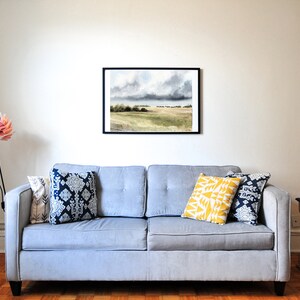 K&ouml;nnte beinhalten: Ein graues Sofa mit drei Kissen, einem gelben, einem blau-wei&szlig;en und einem blau-wei&szlig;en mit geometrischem Muster. Eine schwarze Stehlampe mit Stativ und einem rosa-wei&szlig;en, blumenf&ouml;rmigen Schirm steht in der Ecke des Raumes. Ein schwarzer und goldener Beistelltisch steht rechts vom Sofa. Ein gerahmtes Aquarellbild einer Landschaft mit bew&ouml;lktem Himmel h&auml;ngt an der Wand &uuml;ber dem Sofa.