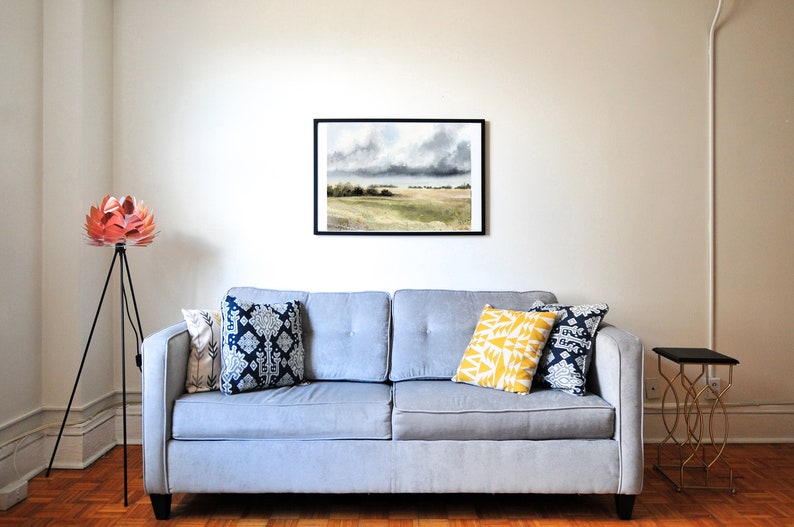 K&ouml;nnte beinhalten: Ein graues Sofa mit drei Kissen, einem gelben, einem blau-wei&szlig;en und einem blau-wei&szlig;en mit geometrischem Muster. Eine schwarze Stehlampe mit Stativ und einem rosa-wei&szlig;en, blumenf&ouml;rmigen Schirm steht in der Ecke des Raumes. Ein schwarzer und goldener Beistelltisch steht rechts vom Sofa. Ein gerahmtes Aquarellbild einer Landschaft mit bew&ouml;lktem Himmel h&auml;ngt an der Wand &uuml;ber dem Sofa.