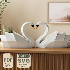 Pode incluir: Duas esculturas de cisnes de papel branco com os seus pescoços formando um formato de coração. Os cisnes têm bicos pretos e detalhes em laranja. A imagem também inclui um ícone de arquivo de corte PDF e SVG.