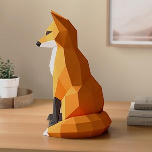 Pode incluir: Uma escultura geom&eacute;trica de raposa laranja com detalhes em preto. A raposa de papel est&aacute; sentada, com a cauda enrolada. A escultura est&aacute; sobre uma superf&iacute;cie de madeira, com plantas e livros ao fundo.