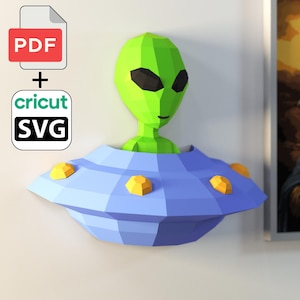Puede incluir: Una cabeza de alienígena de papel geométrico que emerge de un OVNI azul y amarillo. El alienígena es verde con ojos negros. La imagen incluye un icono PDF y Cricut SVG.
