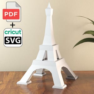 Puede incluir: Una maqueta blanca de la Torre Eiffel de papel se encuentra sobre una superficie de madera. La maqueta está junto a una planta en maceta. La imagen también incluye un icono de archivo PDF y SVG.
