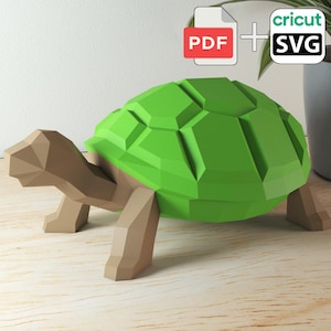 Peut inclure: Une tortue géométrique en papier avec une carapace vert vif et un corps beige. L'image comprend des icônes PDF et SVG, suggérant des fichiers d'artisanat numériques. La tortue est sur une surface en bois.