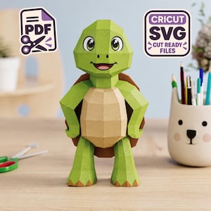 Puede incluir: Una figura de tortuga de papel en 3D con cabeza y extremidades verdes y un plastrón de color tostado. La tortuga tiene ojos grandes y una boca sonriente. Se ven dos iconos: uno etiquetado como PDF con tijeras y otro CRICUT SVG CUT READY FILES.