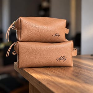 Puede incluir: Dos neceseres rectangulares de color tostado con textura granulada apilados uno sobre otro. Cada bolsa cuenta con un asa lateral, una cremallera con un tirador largo y el nombre Ashley impreso en cursiva negra en la esquina inferior derecha.
