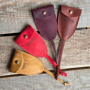 Puede incluir: Cuatro fundas de cuero para llaves en tonos beige, rojo, morado oscuro y marrón, cada una con un cierre de botón a presión de latón y una correa de cuero. Las fundas están grabadas con los nombres Linda, Donna, Vicky y Wendy, dispuestas en abanico.