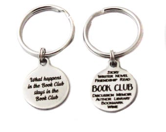 May include: Two silver keychains with round metal tags. One tag says "What happens in the Book Club stays in the Book Club". The other tag says "Story Writer Novel Friendship Read Book Club Discussion Memoir Author Library Bookmark Wine".