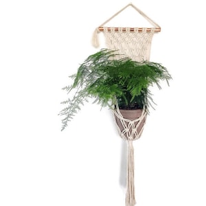 Pode incluir: Um suporte de planta de macramé com uma vareta de madeira e uma franja longa. Uma planta em vaso está pendurada no suporte de macramé.