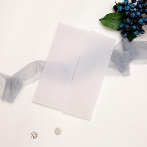 May include: Two white translucent envelopes are displayed on a white surface, alongside a gray tulle ribbon, faux berries, and two decorative rhinestone embellishments. The composition suggests a theme of stationery or wedding invitations.