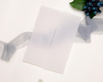 May include: Two white translucent envelopes are displayed on a white surface, alongside a gray tulle ribbon, faux berries, and two decorative rhinestone embellishments. The composition suggests a theme of stationery or wedding invitations.