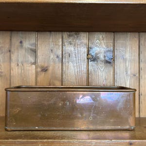 Puede incluir: Una jardinera rectangular de cobre con un acabado metálico pulido y esquinas redondeadas. El recipiente presenta una superficie lisa y reflectante con sutiles variaciones de tono y un diseño abierto en la parte superior.