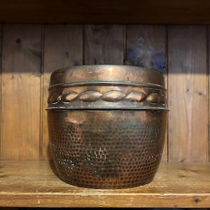 May include: A copper-coloured metal container with a hammered texture and a decorative braided band around the middle. The container sits on a wooden shelf against a wooden plank background.