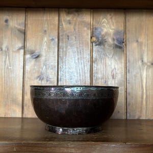 May include: A dark brown, rounded metal bowl featuring a metallic rim and a decorative band with intricate, repeating geometric patterns etched around the upper circumference. The bowl sits on a circular base and has a polished, reflective finish.