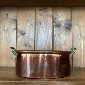 May include: A round copper pot with a polished metallic finish and two curved brass handles attached to the sides. The vessel features a flat base and straight vertical walls, showcasing a warm reddish-brown hue with subtle reflections on its surface.