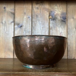 May include: A round copper bowl with a dark, weathered patina and a smooth, curved body. The upper rim features a decorative band with etched geometric patterns, including small crosses and horizontal lines, showing signs of oxidation.