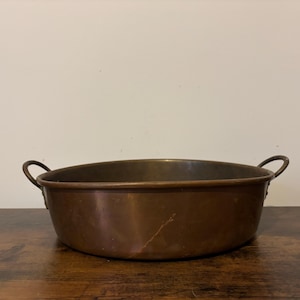 Könnte beinhalten: Eine kupferfarbene, ovale Schüssel mit zwei gebogenen Griffen. Die Metallschüssel steht auf einer Holzoberfläche. Das Innere ist dunkel, und die Außenseite hat eine warme, gealterte Patina. Die Griffe sind klein und gebogen.