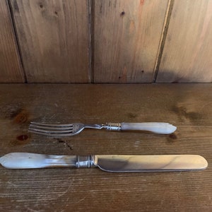 May include: A set of two silver-plated utensils with white pearl handles. The fork and knife are laid out on a wooden surface.