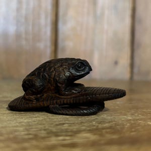 Puede incluir: Una detallada escultura de rana de madera marrón oscuro con detalles texturizados. La rana se asienta sobre una base alargada y texturizada, posiblemente una hoja o un tronco. La escultura está sobre un fondo de madera borroso.