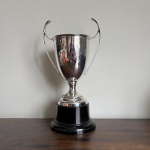 May include: A silver-colored trophy cup with two curved handles, featuring an engraved inscription on the front surface. The cup is mounted on a solid, circular black base, creating a contrast between the metallic finish and the dark stand.