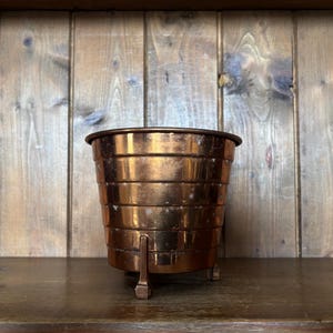 May include: A polished copper planter featuring a tiered, ribbed exterior design with a wide circular opening at the top. The vessel is supported by three small, sturdy metal feet attached to the base, showcasing a metallic finish throughout.