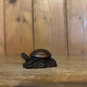 Op de afbeelding: Een kleine, ingewikkeld gesneden houten schildpadfiguur. De schildpad is donkerbruin met gedetailleerde schaalpatronen en zit op een getextureerde basis. Het beeldje staat op een houten oppervlak.
