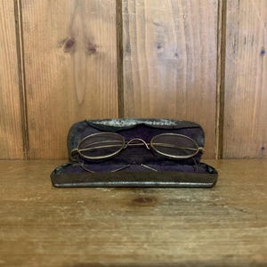 Puede incluir: Un par de gafas redondas con montura dorada en una caja de terciopelo negro con forro morado.