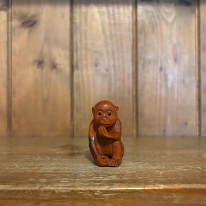 May include: A small, detailed wooden monkey figurine in a seated pose, with its hands clasped near its face. The monkey is a warm brown color and sits on a wooden surface, with a wooden plank background.