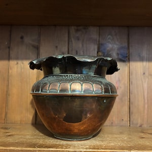 Puede incluir: Un jarrón de metal color cobre con un borde con volantes y un cuerpo bulboso. El jarrón tiene una textura martillada en la parte superior y se encuentra sobre una estantería de madera contra un fondo de paneles de madera. El jarrón mide unos 30 cm de alto.