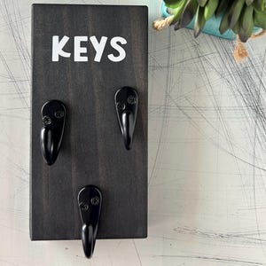 May include: A dark stained wooden key holder with three black metal hooks. The word "KEYS" is printed in white block letters at the top. A decorative succulent plant in a blue pot is in the background.