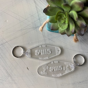 Puede incluir: Dos llaveros de plástico transparente con el texto "De Pere Wisconsin" y el número "54115". Un llavero tiene un diseño floral y el otro es liso.