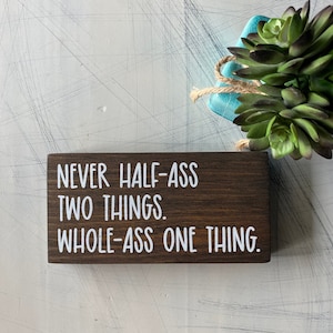 May include: A wooden sign with the text "Never half-ass two things. Whole-ass one thing." in white lettering on a dark brown background.
