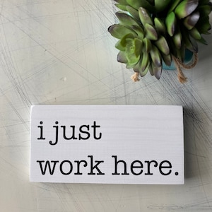 May include: White wooden sign with black text that reads "i just work here."