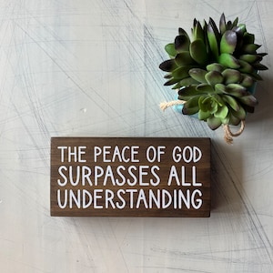 May include: A wooden sign with white text that reads "The peace of God surpasses all understanding". The sign is sitting on a white surface with a small green succulent plant in a blue pot.