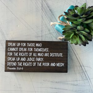 May include: A wooden sign with a white background and black text. The text reads: "Speak up for those who cannot speak for themselves, for the rights of all who are destitute. Speak up and judge fairly: defend the rights of the poor and needy. Proverbs 31:8-9"