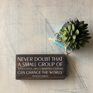 May include: A dark brown wooden sign with white text that reads "NEVER DOUBT THAT A SMALL GROUP OF THOUGHTFUL AND COMMITTED CITIZENS CAN CHANGE THE WORLD. PRESIDENT BARTLET." A small succulent plant in a teal pot is also in the image.