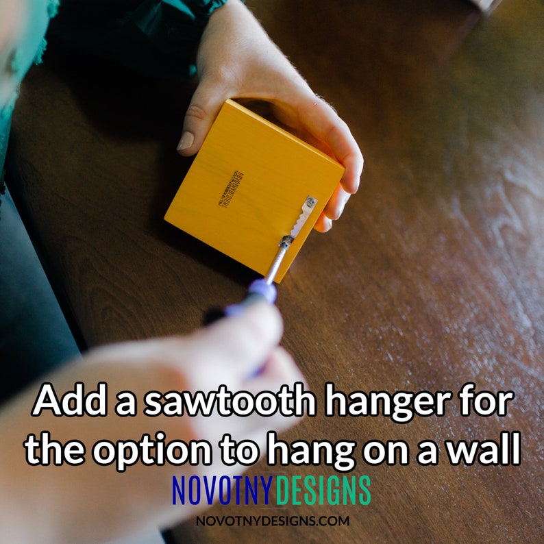 May include: A person is attaching a sawtooth hanger to the back of a yellow wooden sign using a screwdriver. The sign reads "NOVOTNY DESIGNS".