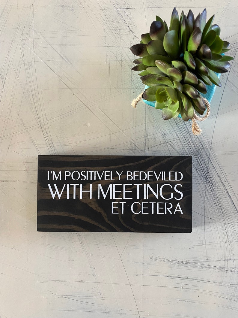 May include: A dark brown wooden sign with white text that reads "I'm positively bedeviled with meetings et cetera".