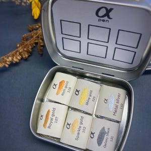 May include: A metal tin with six watercolor paint pans. The pans are labeled with the names of the colors: Royal Gold, Rich Gold, Shiny Gold, Sparkle Gold, Galaxy Black, and Metal Silver. The tin is labeled with the text "α pen".
