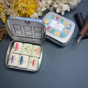 May include: A set of six watercolor paints in a metal tin. The paints are labeled with names like "Sweet Grape", "Strawberry", "Space", "Bright Red", "Sparkle Lizard", and "Tangerine". The tin has a label that says "Summer Limited Collection".