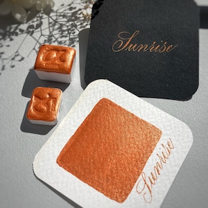 May include: Close-up of watercolor paints in a warm orange hue, labeled "Sunrise." The set includes two square paint cakes and a swatch on a white card. A black tag with "Sunrise" in gold script is also visible.