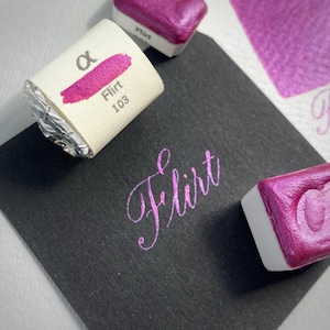 May include: Close-up of watercolor paints and a black surface. The paints are a vibrant magenta color, labeled "Flirt 103." The word "Flirt" is also written in a cursive font on the black surface. The paints are in small, rectangular containers.