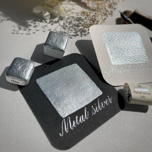 May include: A close-up of a silver watercolor paint pan with the word "Metal Silver" written on a black card. The paint pan is labeled "Metal Silver 110".