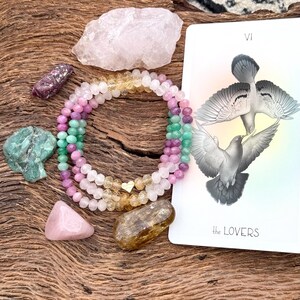 May include: A multi-colored crystal beaded bracelet with a gold heart charm, a pink rose quartz crystal, a green emerald crystal, a brown citrine crystal, a pink rose quartz crystal, and a tarot card with the Lovers card image and the text "VI the LOVERS".