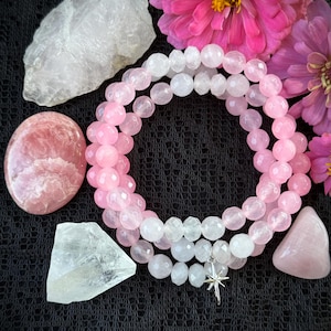 May include: A collection of rose quartz and clear quartz crystals, along with a beaded necklace. The necklace features pink and white beads and a silver star charm. Pink flowers are arranged around the crystals on a black surface.