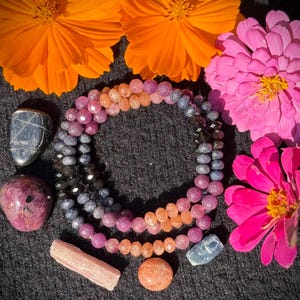 May include: Three beaded bracelets in shades of pink, orange, and blue are displayed with various gemstones and vibrant orange and pink flowers. The bracelets are made of small, faceted beads. The gemstones include a dark blue stone, a pink stone, and a light pink stone.